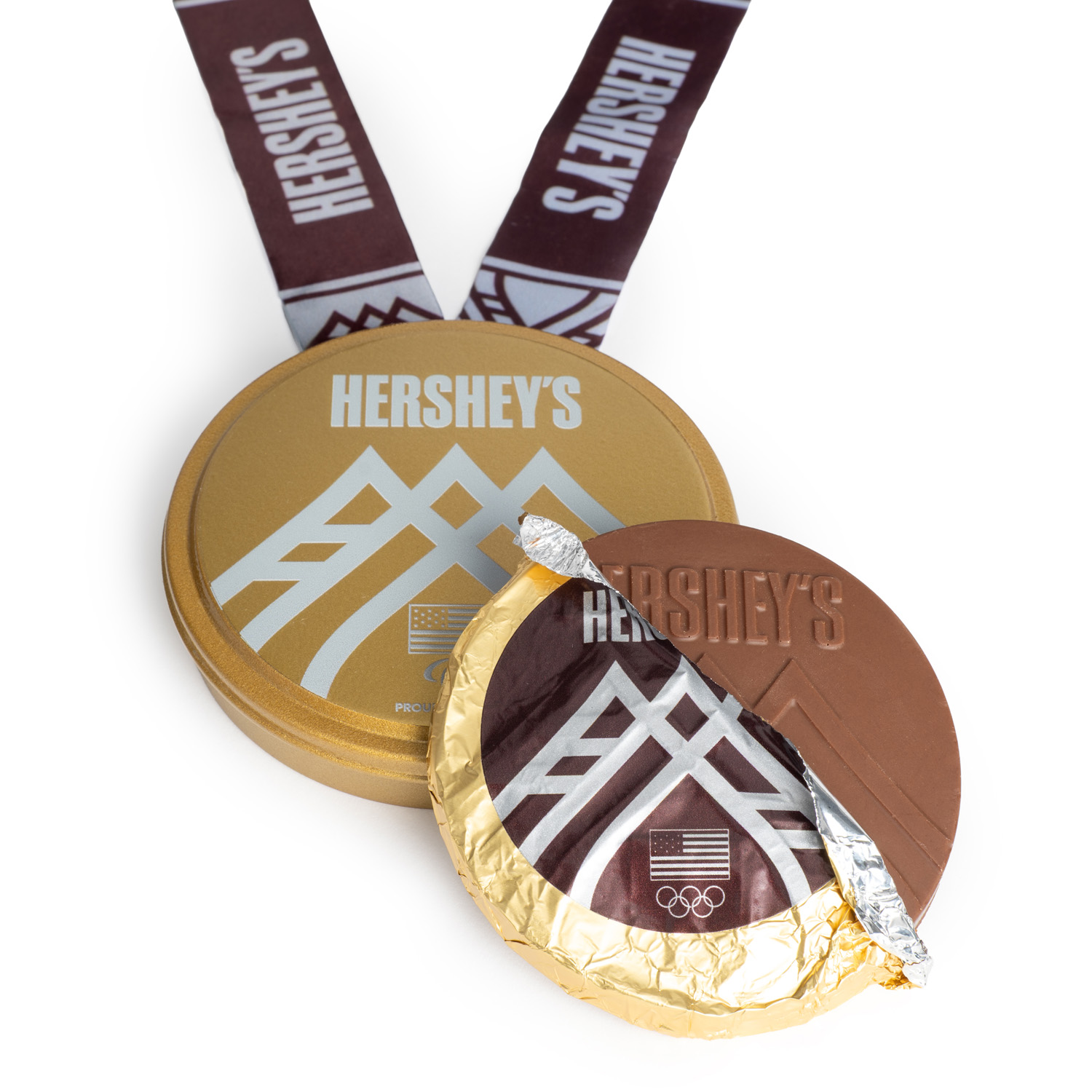 A Hershey’s chocolate “gold medal” partially unwrapped in gold foil sits in front of a round gold tin with a Hershey’s-branded ribbon attached.