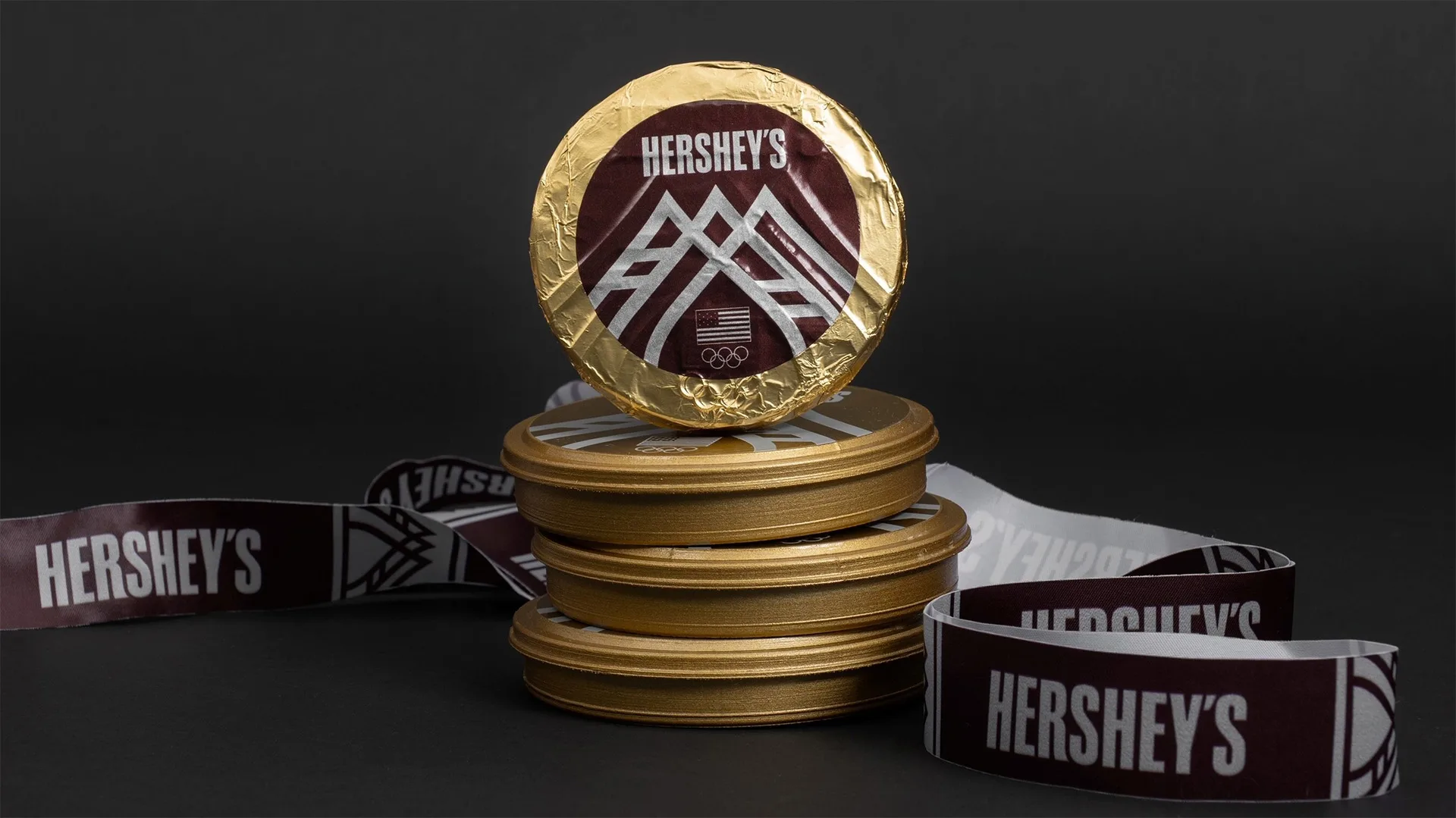 A stack of round Hershey’s gold tins with a chocolate medal displayed on top and branded Hershey’s ribbons arranged around them on a dark background.