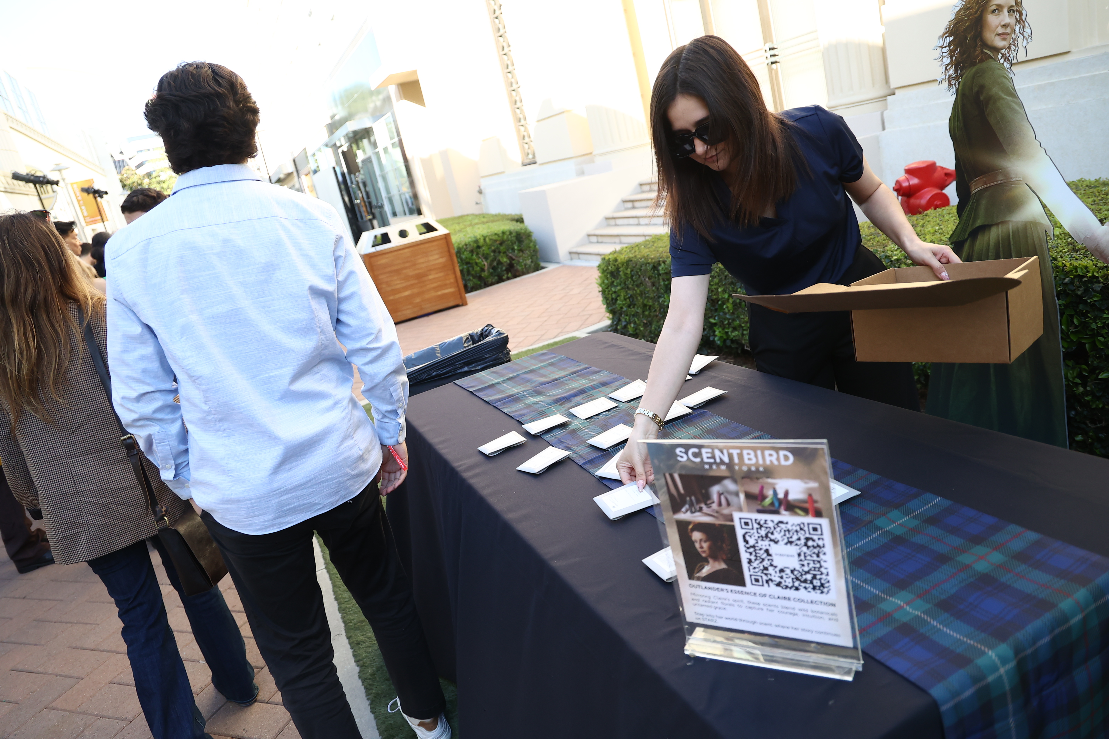 Guests interact with a Scentbird promotional table at an outdoor entertainment event. A staff member wearing sunglasses arranges materials and distributes items from a box while attendees gather nearby. The table features branded signage with a QR code promoting the Outlander-inspired fragrance collection, alongside plaid table décor and character standees in the background.