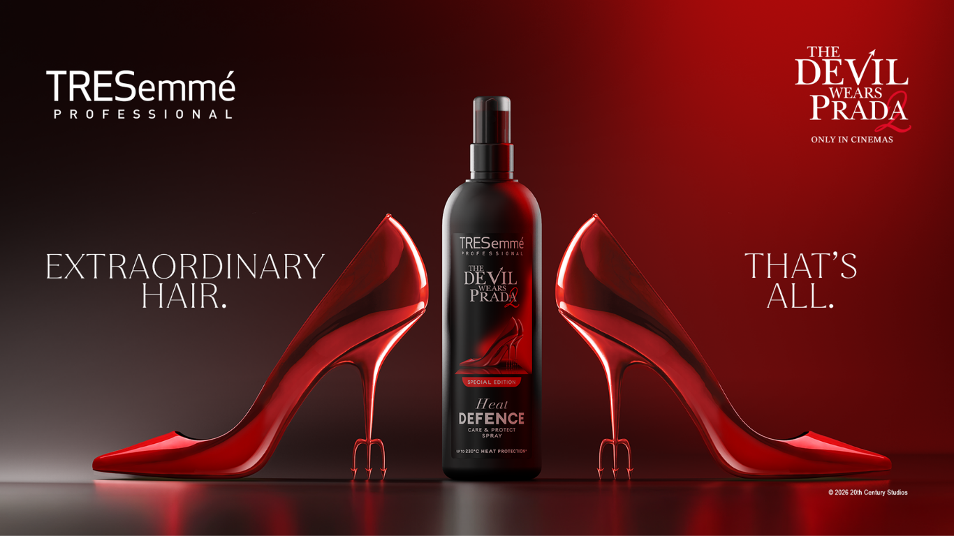 Promotional image for the TRESemmé x The Devil Wears Prada collaboration. A black TRESemmé Heat Defence spray bottle stands centered between two glossy red high heels inspired by the film’s iconic fashion aesthetic. The background features a dramatic red gradient with the TRESemmé Professional logo on the left and The Devil Wears Prada 2 logo on the right, alongside the phrase “Extraordinary Hair. That’s All.”