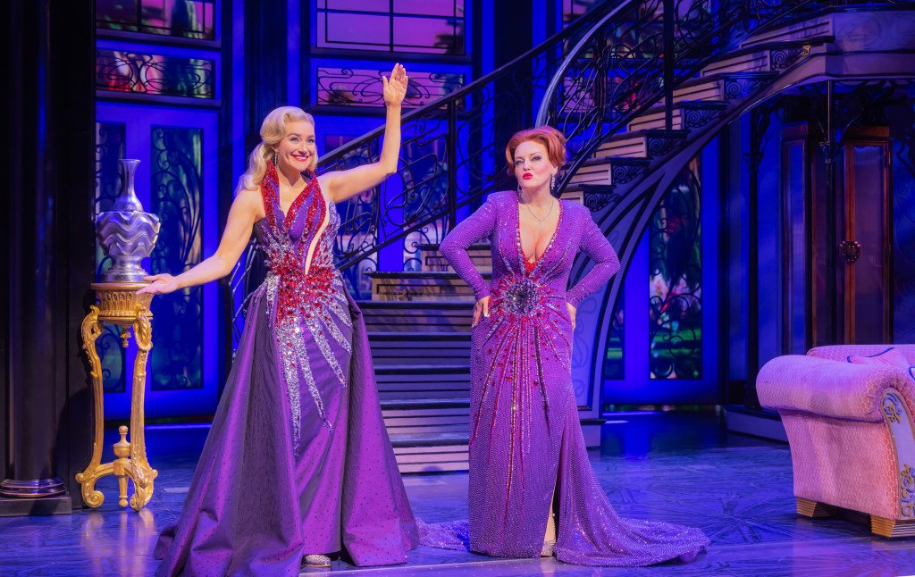 Two performers in sparkling purple gowns stand on a Broadway stage set with dramatic blue lighting, a staircase, and theatrical decor.